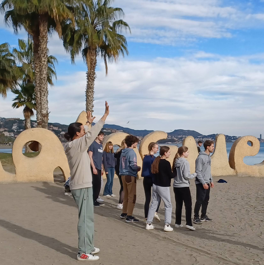 En gruppe elever fra Slotshaven Gymnasium står på stranden med palmer i baggrunden, og kigger mod deres lærer og klassekammerater.