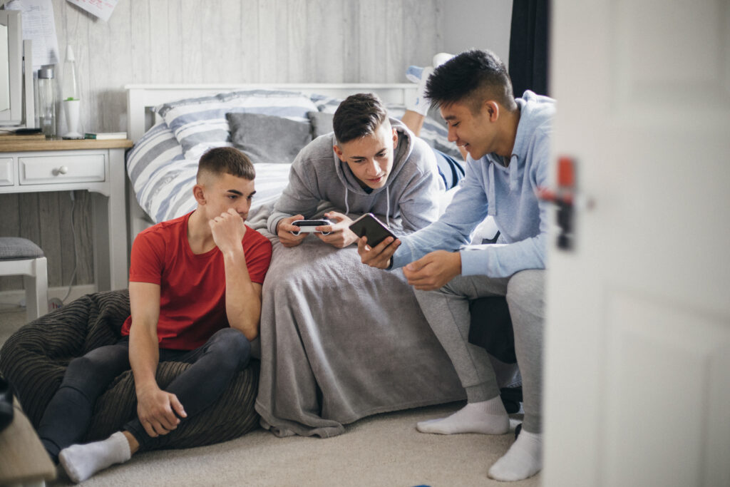 Tre fyre kigger på en telefon. Den ene ligger på en seng, mens en sidder på gulvet og en på en stol.