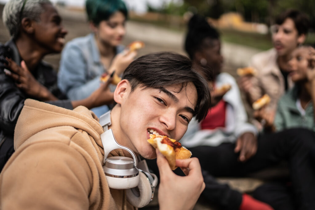 Ung fyr sidder og bidder i et stykke pizza. Hans venner sidder i baggrunden af billedet.