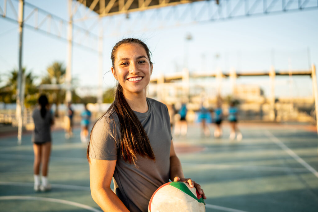 Ung kvinde står med en volleyball i hænderne. I baggrunden kan man se en bane og andre volleyballspillere.