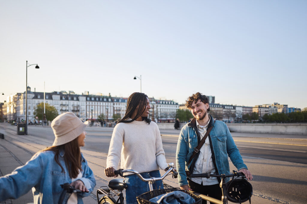 Tre unge går går med deres cykler i København. De kigger på hinanden og smiler, mens de går.