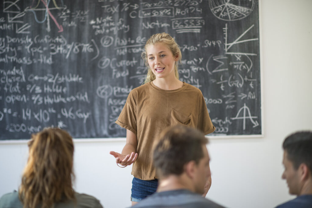 En ung kvinde står og taler foran en lille forsamling på tre, i et klasseværelse.