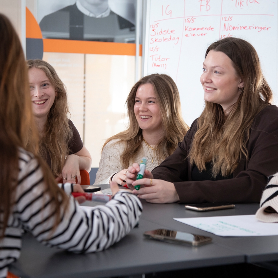 Fire piger fra Slotshaven Gymnasium sidder ved et bord og kigger på en der taler til dem. Pigerne smiler.
