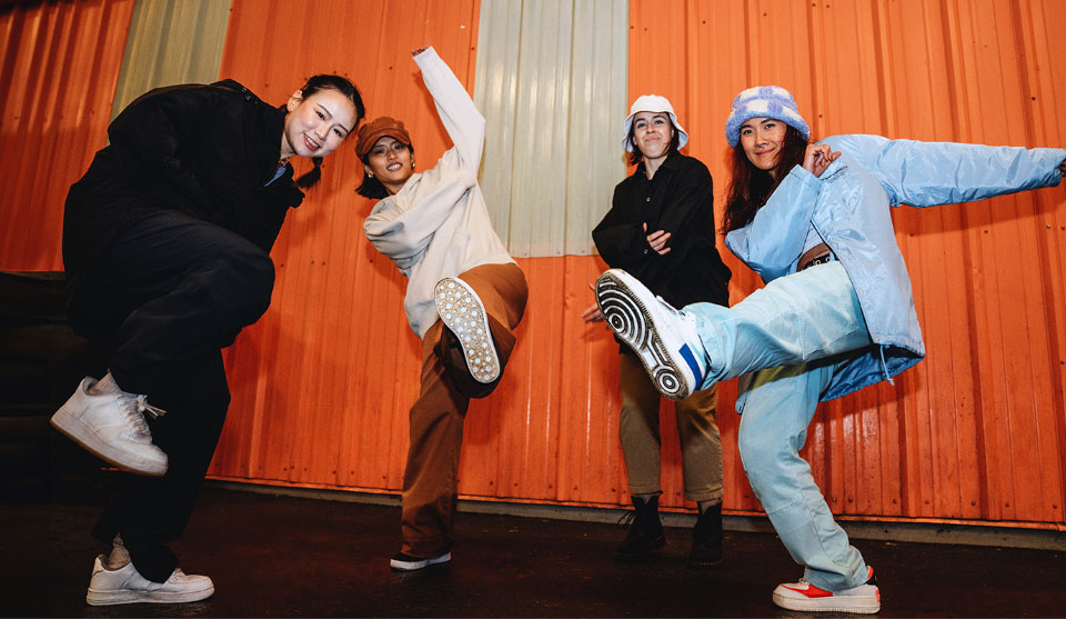 Fire piger danser foran en farvet væg. Pigerne har en hip hop stil.