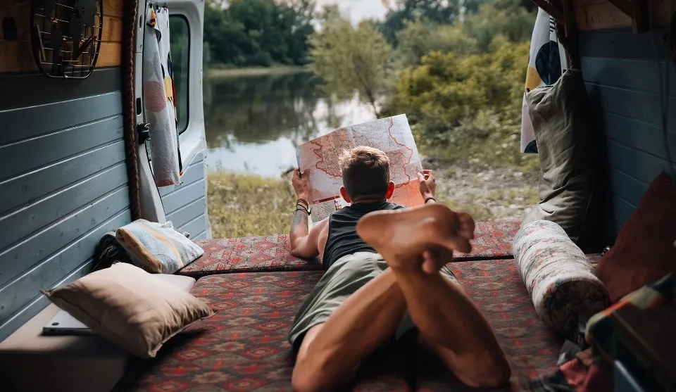 Dreng der ligger på maven i bunden af en varevogn, og kigger på et kort. Der er udsigt til en sø og et grønt område.