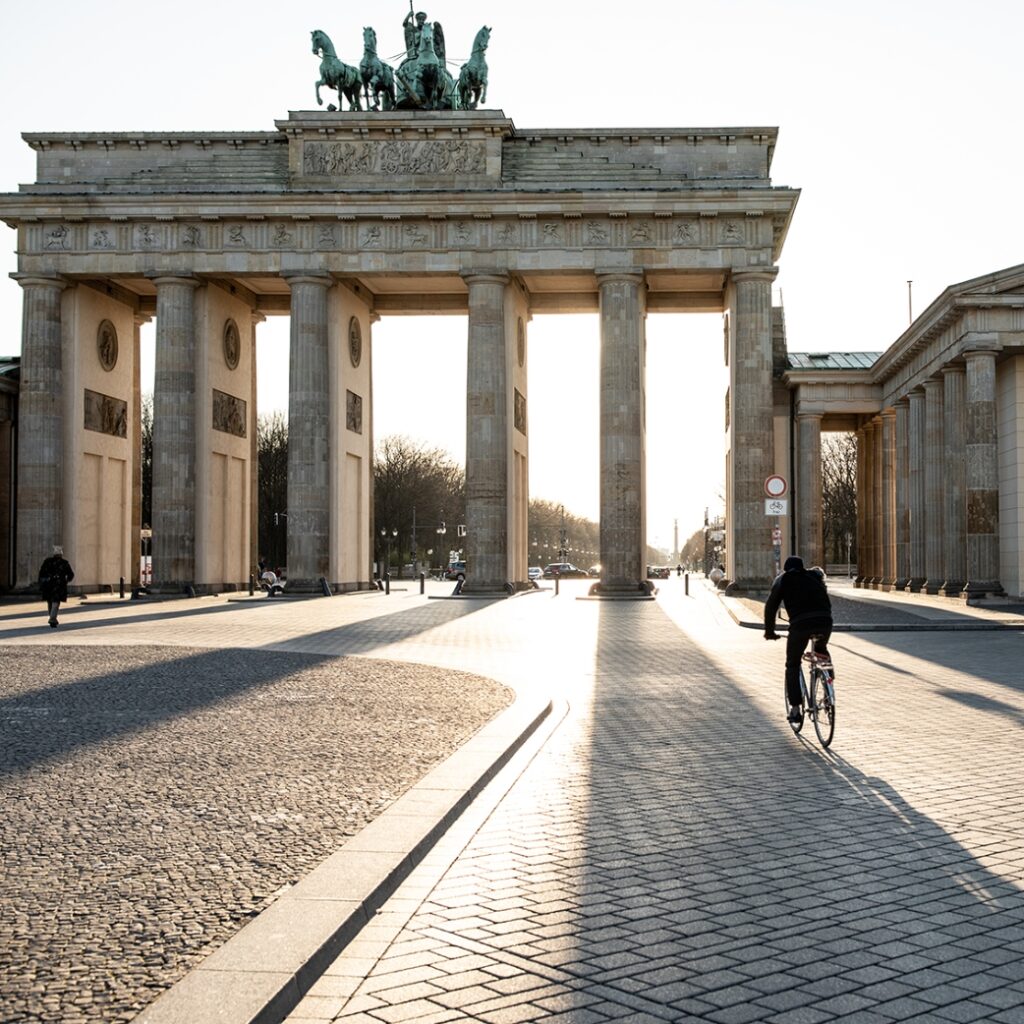 Et billede, taget på afstand af Brandenburger Tor på en solskinsdag. En cykelist cykler hen mod porten.