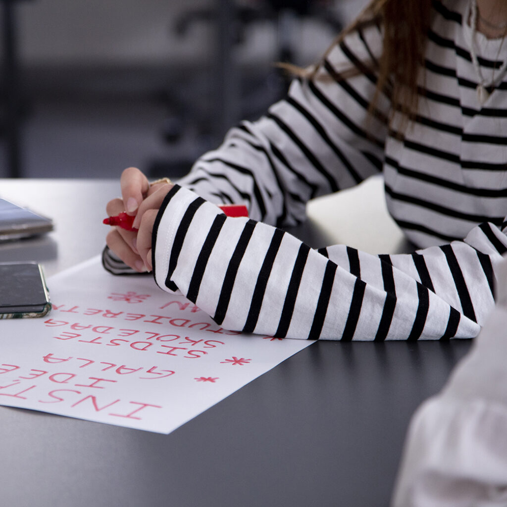 Et billede af en piges arme der læner på et bord, der skriver på et stykke papir med en rød tusch.